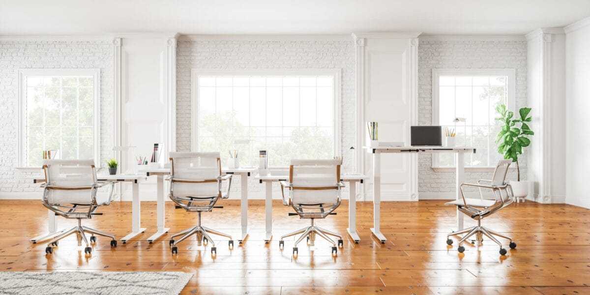Ein heller und moderner Büroraum mit weißen Schreibtischen, ergonomischen Stühlen und großen Fenstern, die natürliches Licht hereinlassen. Der Raum verfügt über weiße Ziegelwände, Holzboden und einen Steh-Schreibtisch-Bereich