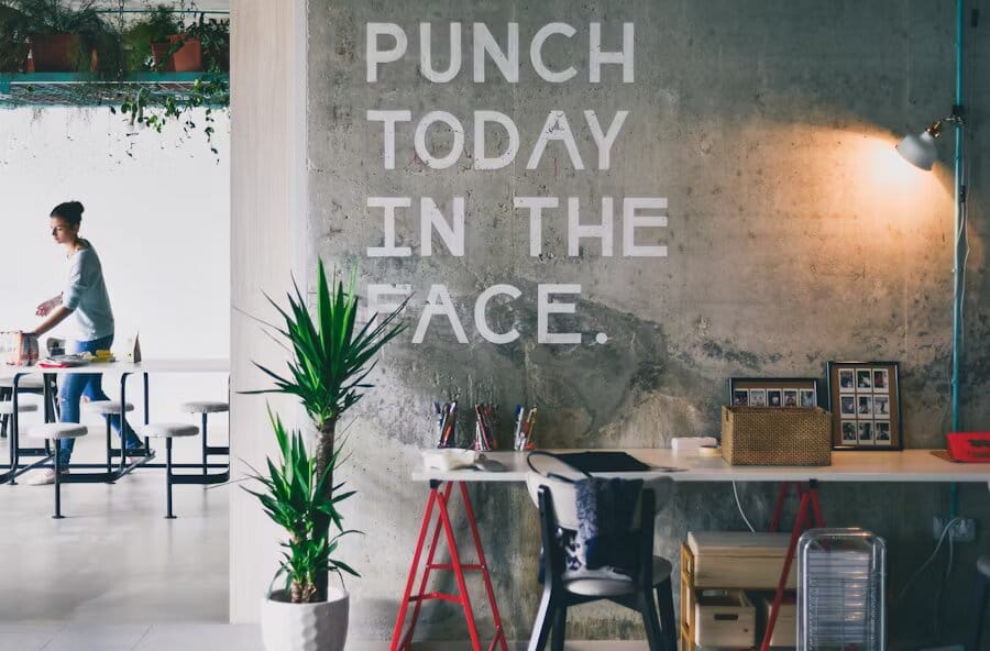 Entspannter Büroraum mit einer Betonwand mit der Aufschrift „Punch today in the face“, grünen Pflanzen, einem weißen Schreibtisch mit Büromaterialien und einer Person im Hintergrund.