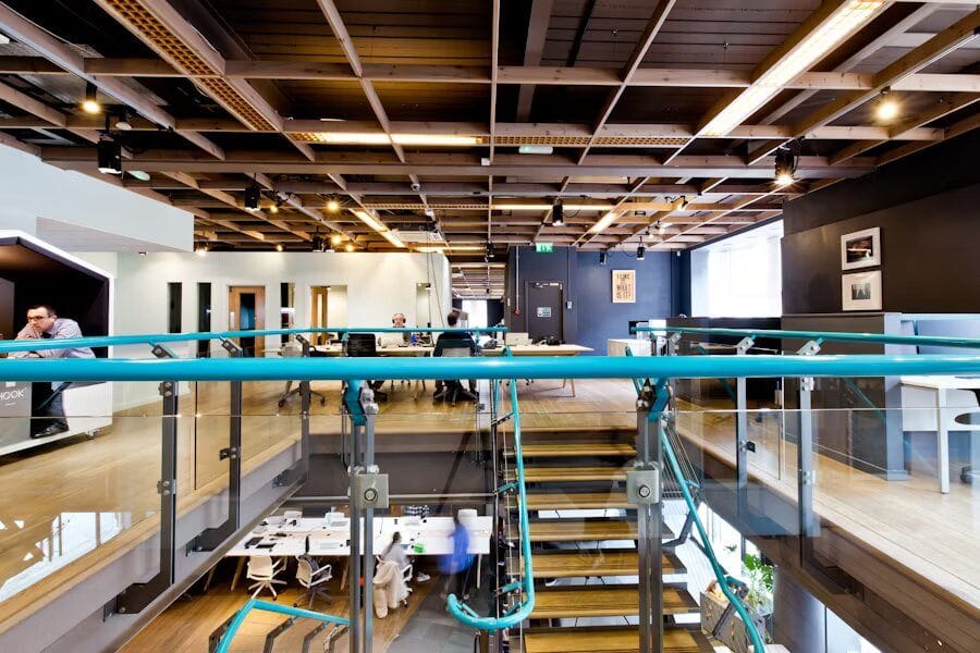 Innenansicht eines modernen Büros mit Zwischengeschoss, offenem Holzdeckensystem, Glastreppe, Coworking-Bereichen und Designermöbeln.