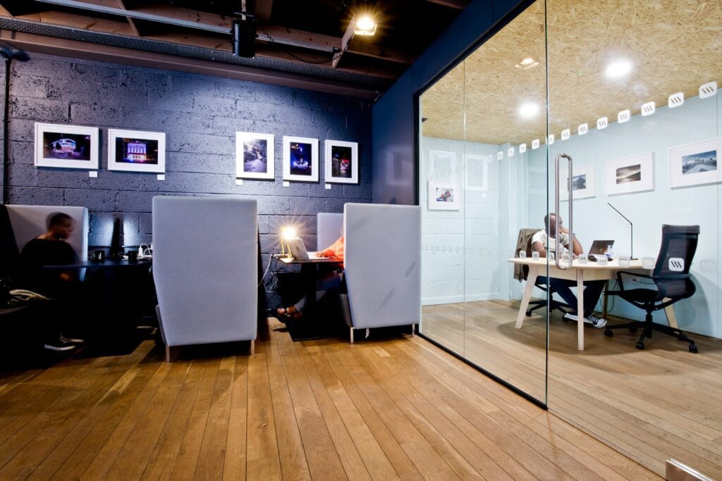 Gemeinsamer Arbeitsbereich mit gemütlichen Arbeitsnischen und einem verglasten Privatbüro, dekoriert mit Fotos an einer schwarzen Wand